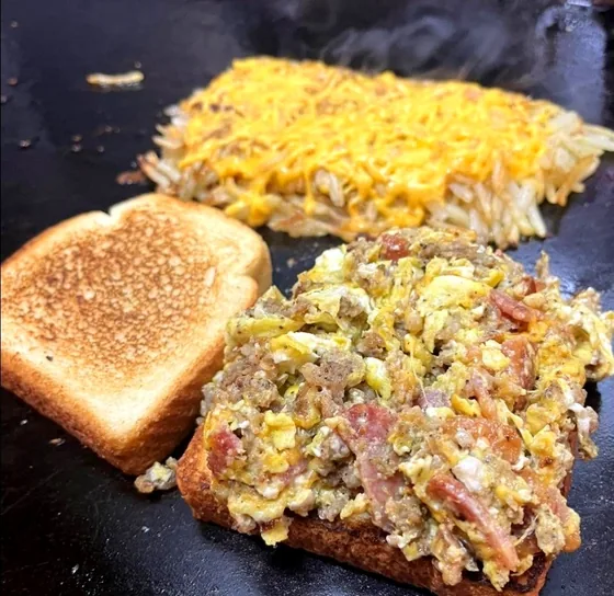 Cheese-covered hashbrown and a loaded scramble on Texas toast cooking on the flat-top